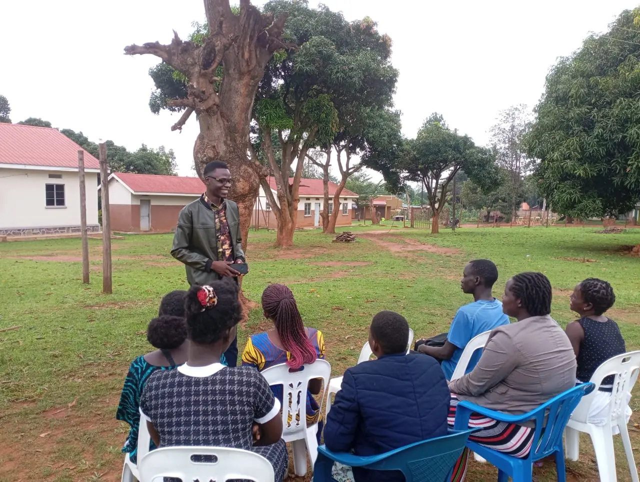 Preacher teaching a seated group of youth and women outdoors under a large tree